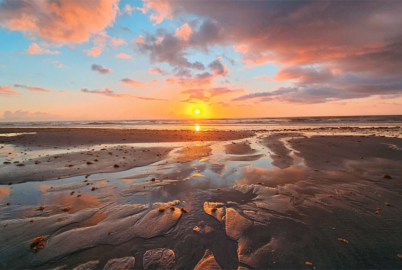 Sunset over a calm sea reflecting orange light on wet sand and shallow tidal pools at the beach.