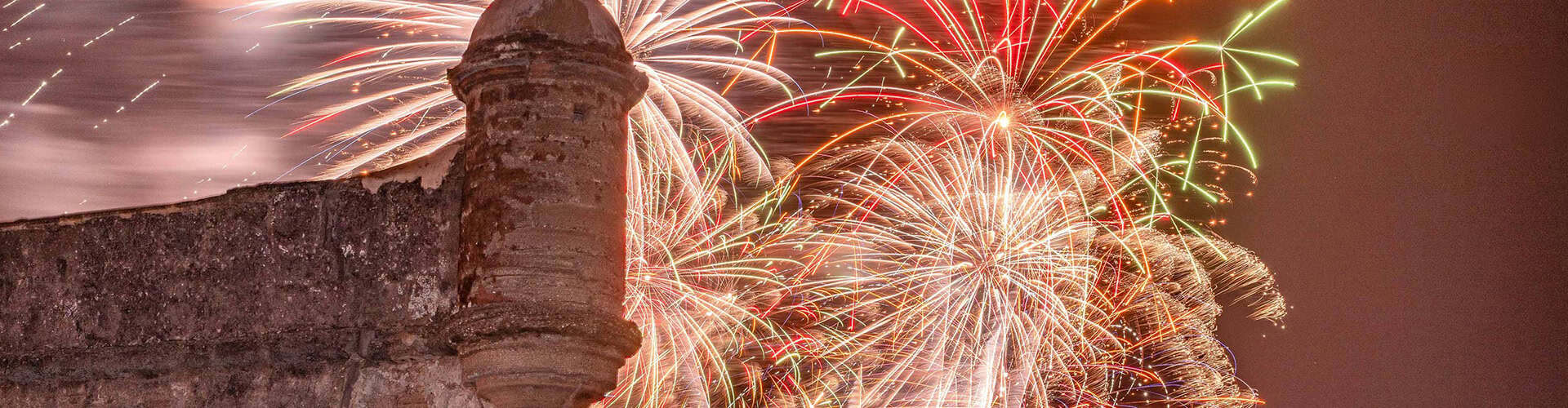 Fireworks explode above a historic stone fortress wall with a rounded turret at night, creating colorful trails in the sky.